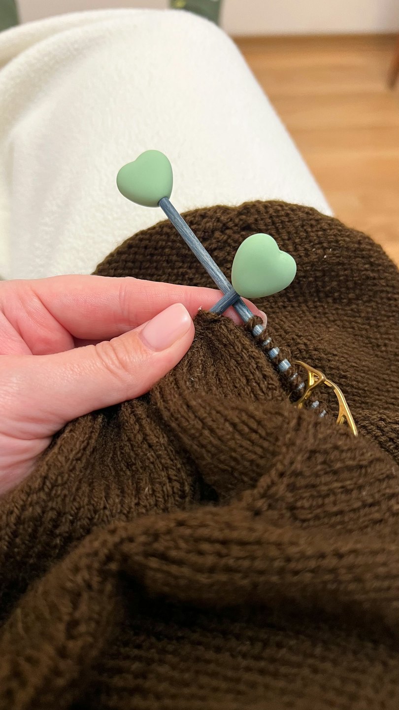 A hand knits brown yarn with a heart-shaped needle.